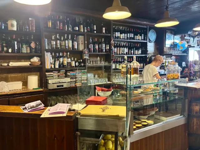 בר ותיק עם מדפי בקבוקים, דלפק עץ, ויטרינה ואישה עובדת מאחוריו