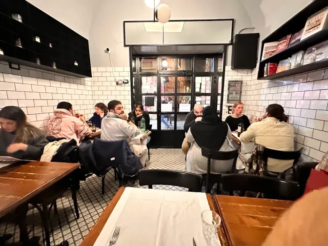 חדר קטן בקירות אריחים לבנים, עמוס סועדים סביב שולחנות כהים