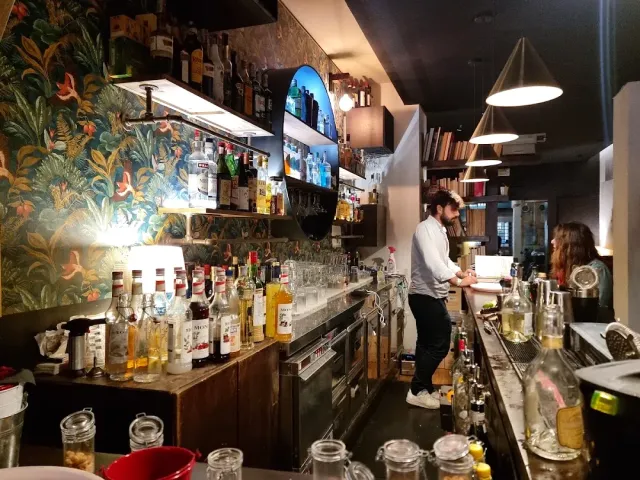 ברמן מאחורי דלפק ארוך, טפט צמחייה צבעוני ומדפי בקבוקים מוארים