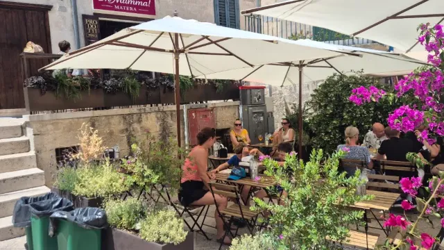 שולחנות עץ תחת שמשיות לבנות בסמטה, סועדים, פרחים סגולים ומדרגות אבן