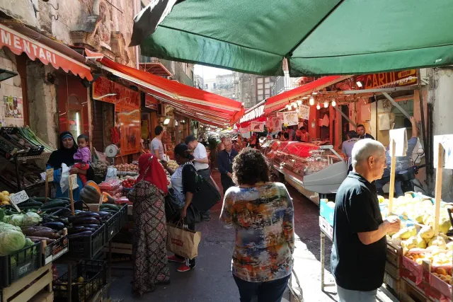 סמטת שוק בין סוככים צבעוניים עם דוכני ירקות וקונים