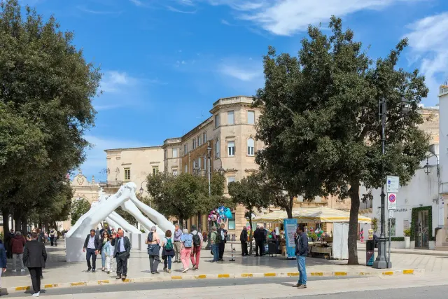 כיכר מרוצפת עם עצים, ספסלים ומבנים לבנים באור יום