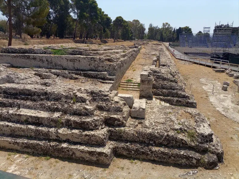 שורות אבן מגולפות של מבנה עתיק חשוף, ועצים ירוקים באופק ביום שמש
