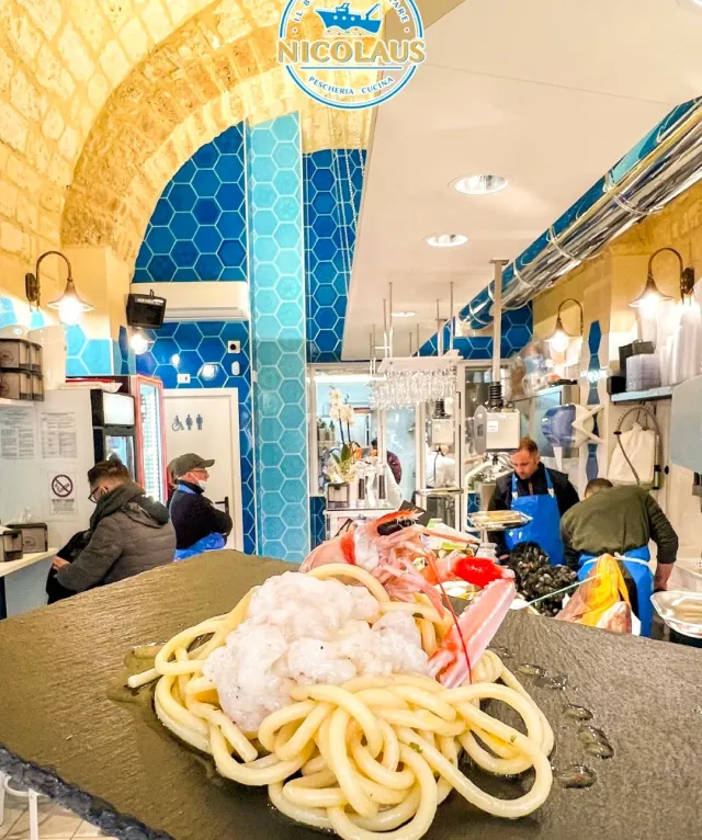 פסטה עם פירות ים טריים על משטח כהה, ומאחור דלפק דגים באריחים כחולים