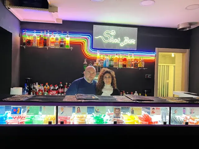 זוג עומד מאחורי דלפק בר עם תאורת ניאון צבעונית ובקבוקים על הקיר
