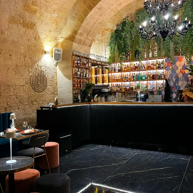 בר במרתף מקומר עם מדפי בקבוקים מוארים, נברשת ורצפת שיש שחורה