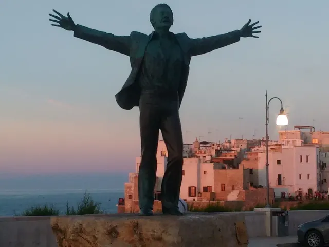 פסל ברונזה של גבר בפרישת ידיים מול עיירה ובים ברקע