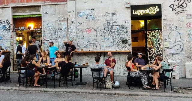 יושבים סביב שולחנות ברחוב לצד קיר מרוסס גרפיטי וכניסה שחורה
