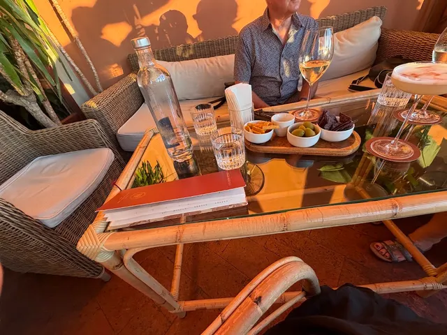 שולחן זכוכית עם יין לבן, זיתים ונשנושים בין כורסאות נצרים באור שקיעה