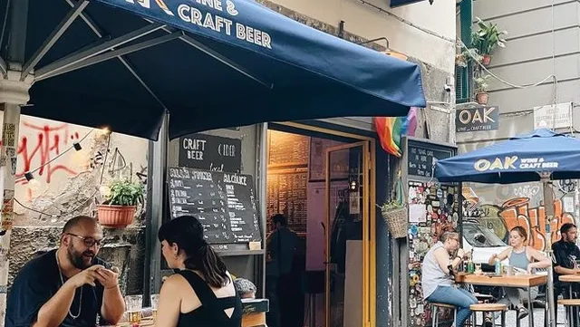 יושבים בשולחנות תחת סוככים כחולים ליד כניסה צרה בסמטה